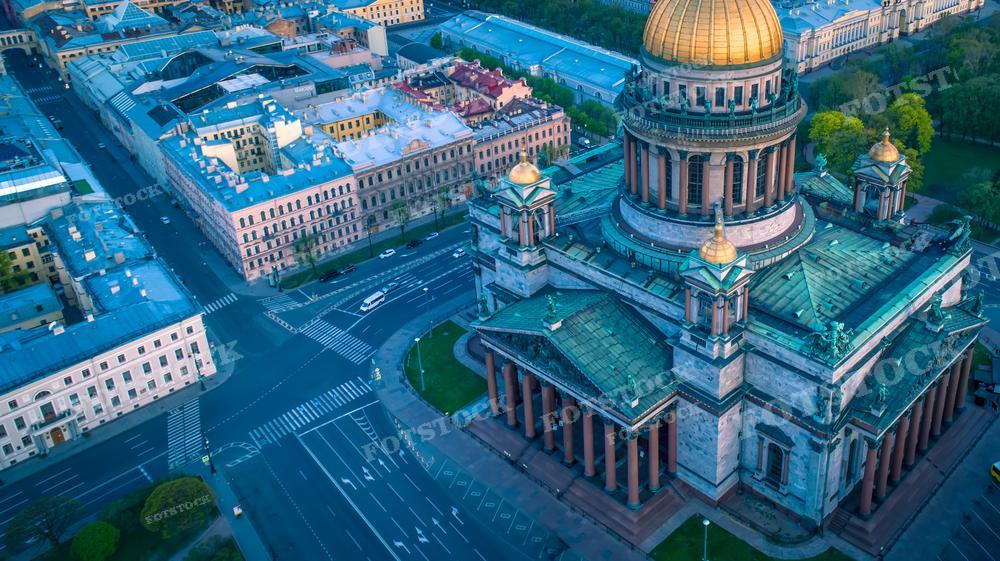 На фотографии запечатлен величественный Исаакиевский собор в Санкт-Петербурге, снятый с воздуха в мягких тонах утреннего или вечернего света. Вокруг собора видна городская застройка, улицы и перекрёстки, добавляющие динамику в статичность архитектурного ансамбля. Этот снимок может быть использован в исторических книгах, туристических буклетах, образовательных материалах и как иллюстрация для статей о русской культуре и православии.